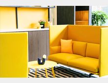 Moderner Innenraum mit einem gelben Sofa mit hohen Seitenlehnen und passenden Kissen, einem runden gelben Tisch mit einer blauen Vase darauf und einem schwarz-weiß gestreiften Teppich. Im Hintergrund eine gelbe Wand mit Holzvertäfelung und dekorativen Pflanzen.