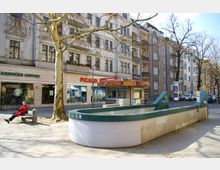 Straßenansicht in einer Stadt mit mehrstöckigen Wohnhäusern im Hintergrund, Geschäften im Erdgeschoss wie einer Starbuck's-Filiale und einer Wurstbraterei. In der Mitte des Bildes steht ein großer Baum, daneben eine Person in roter Kleidung auf einer Bank und eine grün-weiße Brüstung, die zu einem unterirdischen Bereich führt.