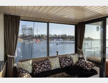 Wohnraum mit großer Fensterfront und Blick auf einen See mit Segelbooten und Stegen. Innenbereich mit Holzbalkendecke, Vorhängen und einer gemütlich dekorierten Sitzbank mit Kissen.