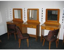 Drei schminke typische Frisier- oder Schminktische aus Holz mit jeweils einem rechteckigen Spiegel und seitlich angebrachten Rundlampen. Vor jedem Tisch steht ein mit Karostoff bezogener Holzstuhl, der Raum hat einen dunklen Teppichboden und eine helle Wand.