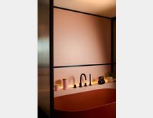 Modernes Badezimmer mit rosa Wänden und einer freistehenden rosa Badewanne. Über der Wanne befinden sich ein schwarzer Wasserhahn, kleine Dekorationsgegenstände, darunter eine schwarze Badeente, und warme, indirekte Beleuchtung.