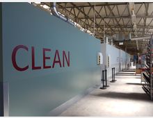 Innenraum einer Industriehalle mit einer hohen Decke aus Stahlträgern. Entlang einer hellblauen Wand mit großen roten Buchstaben "CLEAN" sind Geräte und Absperrungen angebracht. Im Hintergrund sind Regale und Ausrüstungen sichtbar.