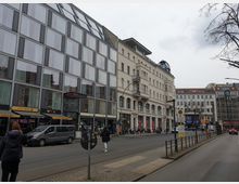 Straßenszene in einer Stadt mit mehrstöckigen Gebäuden, darunter ein modernes Gebäude mit einer Glasfassade links und ein historisches Gebäude mit Kuppel und schmuckvollen Balkonen rechts. Im Vordergrund befinden sich eine Straßenbahnhaltestelle, parkende Autos und Passanten.