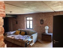 Wohnzimmer in einem industriellen Stil mit freiliegenden Backsteinwänden, Holzboden und einer schlichten Decke. Ein Ecksofa mit Kissen und einer Decke steht nahe einem Fenster, daneben eine rustikale Holztruhe mit einer modernen Lampe darauf.