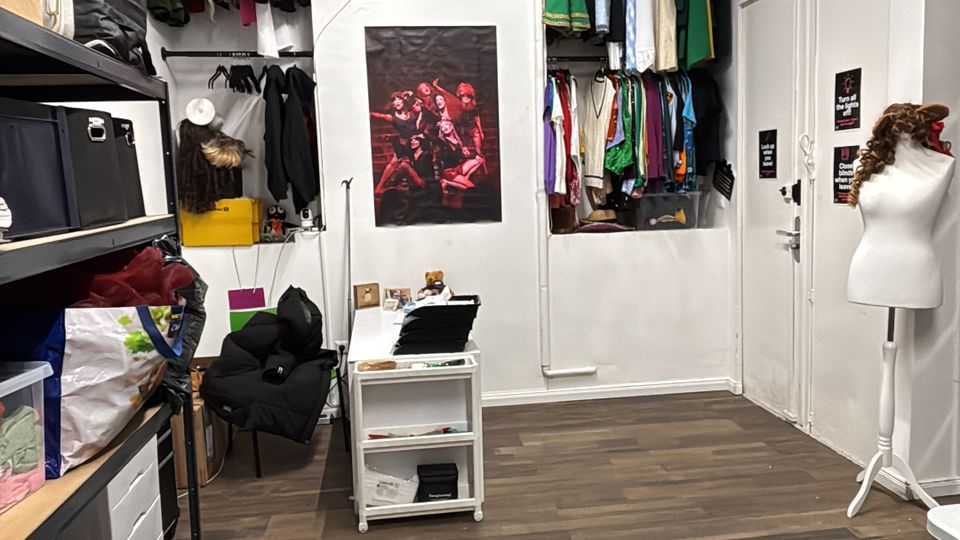 Small costume storage room with white walls and dark wood flooring, featuring shelves of bins on the left and two recessed wall closets with hanging clothes at the back. Overhead exposed pipes and a long fluorescent light run along the ceiling, with a poster on the back wall and a mannequin stand near a set of white doors on the right.