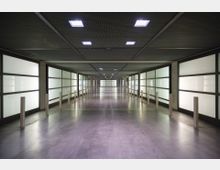 Ein moderner, langer Korridor mit glattem Betonboden und mattem, hinterleuchtetem Glas an den Wänden. Die Decke besteht aus einem Gitterdesign mit integrierten quadratischen Leuchten, und Holzpfosten sind entlang des Weges symmetrisch angebracht.