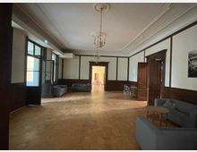 Ein weitläufiger Raum mit Holzboden und Wandvertäfelungen, weißen Wänden und einer dekorativen Decke mit einem Kronleuchter. Der Raum enthält graue Sofas, einen kleinen runden Tisch und Türen, die zu angrenzenden Bereichen führen.