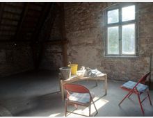 Ein einfacher Dachboden mit unverputzten Backsteinwänden und Holzbalken, erhellt durch ein rechteckiges Fenster. In der Mitte stehen ein Tisch mit Werkzeug und Eimern sowie zwei gepolsterte Klappstühle.