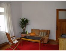 Wohnzimmer mit einfachem Interieur: Eine beigefarbene Ledercouch mit orangefarbenem und gestreiftem Kissen, ein kleiner Holztisch mit schwarzer Platte und zwei Kerzenhaltern, ein Holzstuhl und eine Topfpflanze in einer Ecke. Der Raum hat weiße Wände, einen roten Teppich, helle Vorhänge vor einer Glastür und eine Holztür rechts.