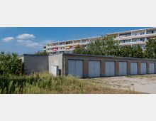 Flache Garagen mit grauen Türen stehen neben einem unbefestigten, bewachsenen Platz, im Hintergrund ein mehrstöckiges Wohngebäude mit Balkonen und bunten Sonnenschirmen, umgeben von Bäumen.