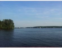 Ein großer See mit ruhiger Wasseroberfläche, mehreren Segelbooten im Hintergrund und einem bewaldeten Ufer auf der gegenüberliegenden Seite. Links im Bild befindet sich ein dichter Baumabschnitt, der ins Wasser hineinragt.