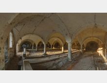 Ehemaliger Stall mit Gewölbedecke und gemauerten Rundbögen, gestützt von massiven Säulen. Der Boden ist mit Stroh und Schmutz bedeckt, die Wände sind teils aus freiliegendem Ziegelstein, und durch Fenster fällt gedämpftes Licht.