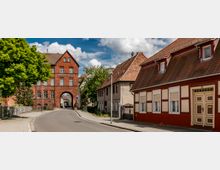 Straßenszene in einer Kleinstadt mit dem markanten Backsteingebäude des Spreewald-Museums im Hintergrund, das einen Torbogen hat, durch den eine schmale Straße führt. Links und rechts stehen Häuser mit roten Ziegeldächern, einige Fachwerk-Elemente sind sichtbar, und ein Baum steht am Straßenrand.