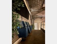 Reihe von blauen Kinositzen in einem industriellen Raum mit freiliegenden Ziegelwänden und einer Decke aus Beton und Metallträgern; links und rechts sind dekorative grüne Pflanzen zu sehen, im Hintergrund eine geschlossene weiße Tür.