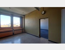 Ein leerer Innenraum mit einem Fenster, durch das ein Blick auf industrielle Gebäude im Hintergrund sichtbar ist. Der Raum hat gelbbraune Wände, einen abgenutzten Bodenbelag, eine analoge Wanduhr und eine Türöffnung, die in einen angrenzenden Bereich mit blauem Teppichboden führt.