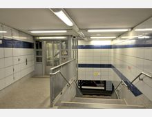 Unterirdischer U-Bahnhof mit weiß gekachelten Wänden, ergänzt durch blaue Akzente. Eine Treppe mit Handläufen führt hinunter, während ein Aufzug und ein Schild mit der Beschriftung „U5 Alexanderplatz ↔ Hönow“ sichtbar sind.