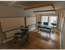 Ein schallisolierter Musikraum mit einem elektronischen Schlagzeug links, einer Arbeitsstation mit Monitor, Tastatur und Lautsprechern rechts sowie Holzfußboden. Die Wände und Decke sind mit Akustikplatten verkleidet, ein Fenster bietet Ausblick auf ein benachbartes Gebäude.