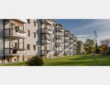 Mehrstöckiges Wohngebäude mit mehreren Balkonen, umgeben von einer großzügigen Grünfläche mit vereinzelten Bäumen. Im Hintergrund sind parkende Autos und weitere Vegetation sichtbar.