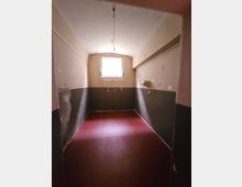 Ein kleiner, schlichter Raum mit roten Bodenfliesen und hellen Wänden, die im unteren Bereich dunkel gestrichen sind. Am Ende des Raums befindet sich ein kleines Fenster, das Tageslicht hereinlässt, und an der Decke hängt eine lose Lampe.