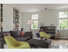 Wohnzimmer mit heller Einrichtung, bestehend aus einer großen grauen Sofalandschaft, einem limettengrünen Sessel, einem dunklen Holzkavier und zwei hohen Fenstern mit Speichenrahmen. Im Hintergrund sind weiße Bücherregale und eine Stehleuchte sichtbar, der Boden besteht aus hellem Holzparkett.