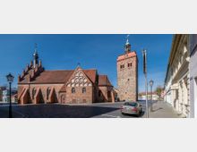 Straßenszene mit einer historischen Backsteinkirche und einem massiven, angrenzenden Glockenturm in romanisch-gotischem Stil. Im Vordergrund befinden sich Kopfsteinpflaster, eine Straßenlaterne und ein geparktes Auto; rechts sind umliegende Gebäude sichtbar.