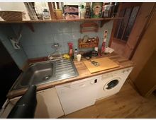 Kleine Küche mit Spüle, Holzarbeitstisch und blauen Fliesen an der Wand. Unter der Arbeitsplatte befinden sich eine Waschmaschine und eine Spülmaschine; darüber ein Regal mit Gewürzdosen, Gläsern und Küchenutensilien.