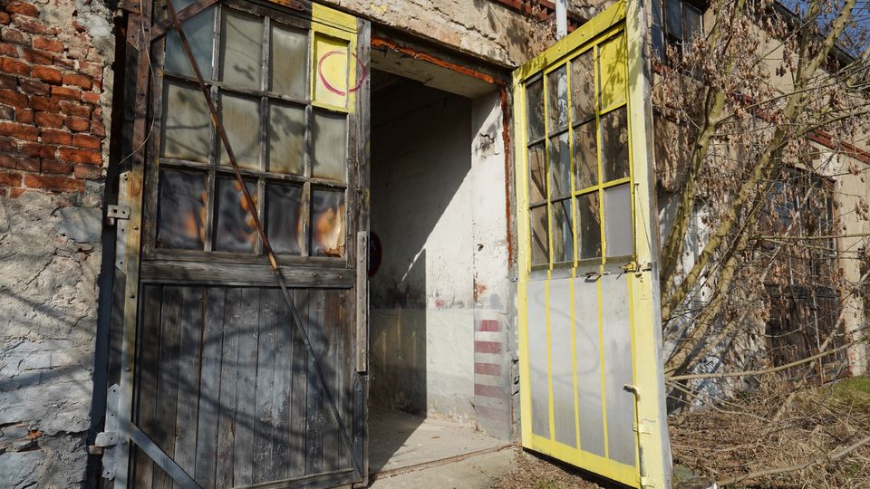 Eingang eines verfallenen Backstein- und Putzgebäudes mit zwei geöffneten, verglasten Holztüren, eine dunkel und eine gelb gestrichen. Vor dem Eingang stehen trockene Sträucher und Äste, der Boden ist teils Beton, teils Gras.