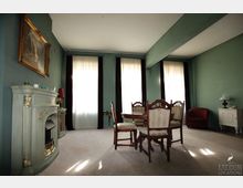 Wohnzimmer mit grünen Wänden, einem dekorativen Kamin, einem runden Esstisch aus Holz mit gepolsterten Stühlen und großen Fenstern mit weißen Vorhängen und dunklen Vorhängen. Im Raum sind außerdem ein roter Sessel, eine weiße Kommode und ein gerahmtes Bild über dem Kamin zu sehen.