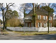 Eckgrundstück mit einem zweistöckigen Backsteinhaus im klassischen Stil, weißen Fensterläden und einem markanten runden Vorbau. Das Grundstück ist von einem weißen Lattenzaun umgeben, im Hintergrund stehen Bäume und Gebüsch.