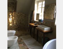 Ein Badezimmer mit einem Waschtisch aus Holz, einem rechteckigen weißen Waschbecken und einem Wandspiegel. Links befinden sich eine offene Dusche mit beigefarbenen Fliesen und ein Bidet sowie eine Toilette, der Boden ist aus Steinfliesen, und Tageslicht fällt durch ein großes Fenster ein.