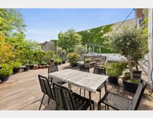 Große Terrasse mit Holzdielenboden, umgeben von verschiedenen Pflanzen in Töpfen. Im Vordergrund steht ein Esstisch mit schwarzen Stühlen, im Hintergrund befinden sich Sitzmöbel und eine Wand, die mit Efeu bewachsen ist.