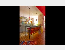 Eine moderne Küche mit einer Arbeitsinsel aus Metall und Glas, die orangefarbene Beleuchtung hervorhebt. Im Hintergrund befindet sich eine Wand mit farbigen Glasfenstern und grauen Schränken, der Boden besteht aus Holz.