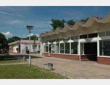 Straßenansicht eines einstöckigen Gebäudekomplexes mit Wellenform-Dach, großen Glasfronten und roten Klinkerwänden. Im Vordergrund ein gepflasterter Gehweg, ein Grünstreifen und ein einzelner Laternenmast, umgeben von Bäumen und klarem Himmel.