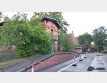 Backsteingebäude im historischen Stil mit Satteldächern, umgeben von Bäumen und Büschen. Eine gepflasterte Zufahrt mit Absperrpfosten führt entlang der roten Ziegelmauer.