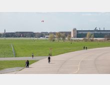Weitläufige Parklandschaft auf dem ehemaligen Flughafengelände Tempelhofer Feld in Berlin, mit großen Rasenflächen, Spaziergängern, Radfahrern und dem ehemaligen Flughafengebäude im Hintergrund. Am Himmel sind bunte Drachen zu sehen, im Hintergrund erkennt man die städtische Umgebung.