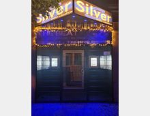 Eingang einer Event-Location bei Nacht mit großem, beleuchtetem „Silver“-Schriftzug und Lichterkette unter dem Vordach. Vor dem Eingang befindet sich ein Metallgittertor; dahinter sind beleuchtete Fenster und ein Banner mit Veranstaltungsankündigung zu sehen.