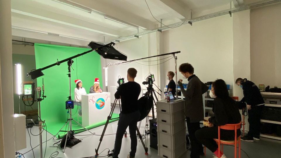 Innenansicht eines Filmstudios mit großer Greenscreen-Wand, Studiobeleuchtung, Kamerastativen und zahlreichen Kabeln. Im Raum stehen außerdem ein weißes Podest vor dem Greenscreen sowie Technik- und Arbeitsbereiche mit Möbeln und Geräten.
