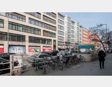 Straßenansicht einer urbanen Umgebung mit mehrstöckigen Geschäftsgebäuden entlang der Straße, geparkten Autos, Fahrrädern vor einem Fahrradständer und einem Eingang zur U-Bahn-Station "Bismarckstraße". Eine Person läuft auf dem Bürgersteig, und im Hintergrund sind Bäume und Werbeschilder sichtbar.