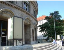 Gebäudeecke aus hellem Stein mit runder Fassade, verziert mit floralen Reliefs oberhalb der Fenster. Im Vordergrund befinden sich halbrunde Treppenstufen mit einem geländerlosen Podest sowie eine leuchtkastenähnliche Konstruktion; im Hintergrund stehen Bäume und Gebäude mit roten Ziegeldächern.