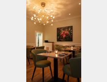 Ein kleiner, eleganter Restaurantraum mit Holzboden, grün gepolsterten Stühlen und hellen Holztischen, die mit Kerzenlicht beleuchtet sind. An der Decke hängt ein moderner Kronleuchter, und an der Wand ist ein Gemälde mit rosa Flamingos zu sehen.