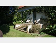 Hausfassade mit weißer Wand und grünen Rankpflanzen, eine kleine Terrasse mit steinernen Stufen und zwei Hundeskulpturen. Im Vordergrund ein gepflegter Garten mit Rasen, Sträuchern und Blumen.