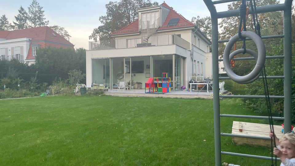 Blick über einen großen Rasen auf ein modernes, weißes Wohnhaus mit großen Glasfronten, Terrasse und Dachterrasse unter rotem Satteldach. Rechts im Vordergrund steht ein Spielgerüst mit Leiter und hängenden Turnringen, umgeben von Bäumen und weiteren Häusern im Hintergrund.