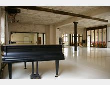 Ein heller, großzügiger Raum mit industriellem Charakter, gekennzeichnet durch freiliegende Betonwände, große Spiegel, Metallpfeiler und eine glänzende weiße Bodenfläche. Im Vordergrund steht ein schwarzer Flügel, während im Hintergrund Fenster und Glaswände zu erkennen sind.