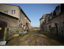 Verlassener Hofdurchgang zwischen zwei alten, stark verwitterten Wohngebäuden mit bröckelndem Putz und teils zugemauerten Fenstern. Ein schmaler, unebener Pflasterweg führt durch ein offenes Metalltor hinaus in eine grasige Freifläche unter blauem Himmel.