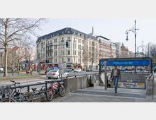 Straßenszene mit dem U-Bahneingang "Adenauerplatz" in Berlin, umgeben von mehrstöckigen Wohn- und Geschäftshäusern mit historischer und moderner Architektur. Im Vordergrund Fahrräder an einem Geländer, Autos auf der Straße und Passanten in Bewegung.