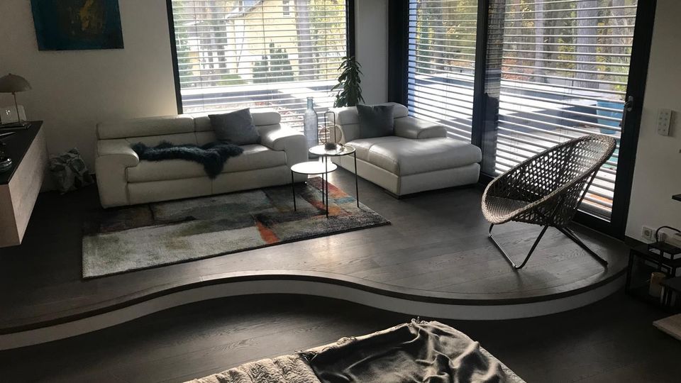 Modernes Wohnzimmer mit heller Eckcouch auf einem Podest, kleinem Rundtisch und Teppich vor großen Fenstern und einer Terrassentür mit Jalousien. Daneben stehen ein geflochtener Lounge-Sessel und eine Zimmerpflanze; draußen ist eine Holzterrasse mit Blick ins Grüne zu sehen.
