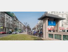 U-Bahnhof-Eingang Theodor-Heuss-Platz in einer städtischen Umgebung mit mehrstöckigen Wohn- und Geschäftshäusern im Hintergrund; Fahrräder sind an einem Ständer abgestellt, und Autos fahren auf der Straße.