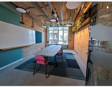 Moderner Besprechungsraum mit Backsteinwänden, einem ovalen Tisch mit bunten Stühlen und einer großen Fensterfront, die Tageslicht einlässt. An der linken Wand befinden sich eine weiße Schreibtafel und eine grüne Akzentfläche, die rechte Wand ist mit hängendem Grün dekoriert.