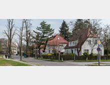 Straßenszene in einer Wohngegend mit Einfamilienhäusern im traditionellen Stil, weißen Fassaden und roten Ziegeldächern. Bäume und Hecken säumen die Straße, auf der Verkehrszeichen und Laternenpfähle stehen; im Hintergrund sind moderne Gebäude sichtbar.