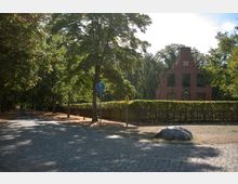 Pflasterstraße in einer von Bäumen gesäumten Umgebung mit einem roten Backsteingebäude im Hintergrund. Vor dem Gebäude befindet sich eine niedrige Hecke und ein Holzzaun, daneben stehen ein Radweg-Schild und ein großer Stein auf der rechten Straßenseite.